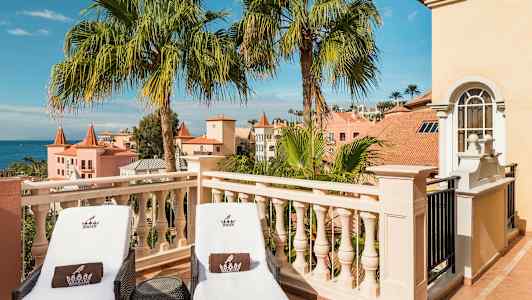 To liggestole på en terrasse med udsigt over palmer og pastelfarvede bygninger. (AI-genereret tekst)