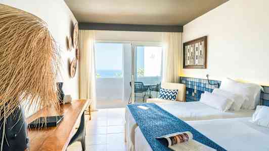 Et lyst hotelværelse med to senge, en træbænk og en altan med havudsigt. (AI-genereret tekst)