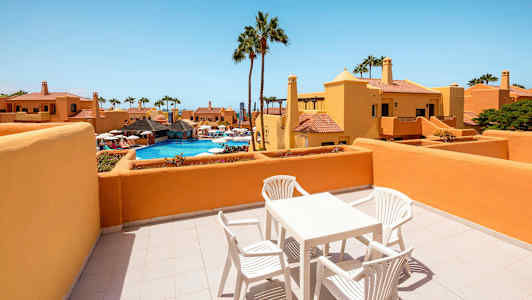 En terrasse med bord og stole, med udsigt over en pool og omkringliggende bygninger. (AI-genereret tekst)