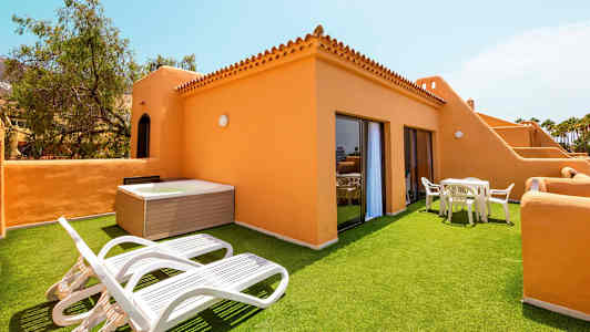 Terrasse med kunstgræs, liggestole, bord og stole samt en jacuzzi, omgivet af orange bygninger. (AI-genereret tekst)