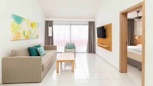 Et hotelværelse med en delvist synlig seng, en sofa og en lænestol samt et bord. Et vindue med gardiner er synligt i baggrunden. (AI-genereret tekst)