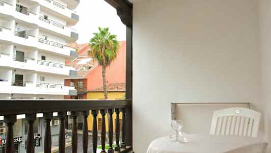 En altan med et bord, to glas og en hvid plaststol. Udsigten viser bygninger, en palme og en gade. (AI-genereret tekst)