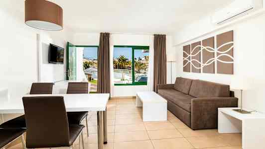 Et lyst og moderne indrettet hotelværelse med en brun sofa, et spisebord og stole. Vinduer og dør leder til udendørsområdet. (AI-genereret tekst)