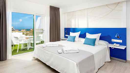 Billede af et hotelværelse med en dobbeltseng, blå rygstøtte og et vindue, der viser en balkon med møbler og udsigt til havet. (AI-genereret tekst)