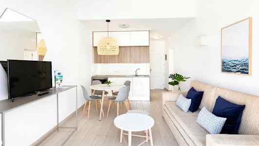 En moderne stue med en beige sofa, blå puder, et hvidt rundt sofabord og et TV på et stativ. Ved siden af er der et spiseområde med et rundt bord og grå stole samt et køkken i lyse farver. (AI-genereret tekst)