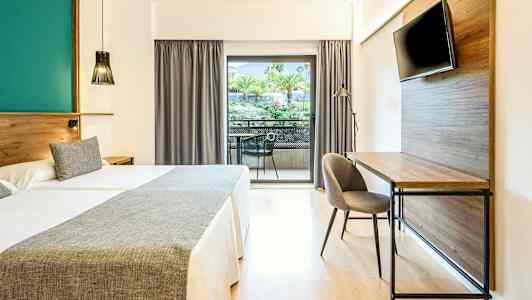 Hotelværelse med to senge, skrivebord, tv og balkon med udsigt over træer og grønne områder. (AI-genereret tekst)