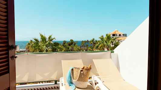 Udsigt over en solrig altan med liggestole, en kurvtaske, palmer og havet i baggrunden. (AI-genereret tekst)