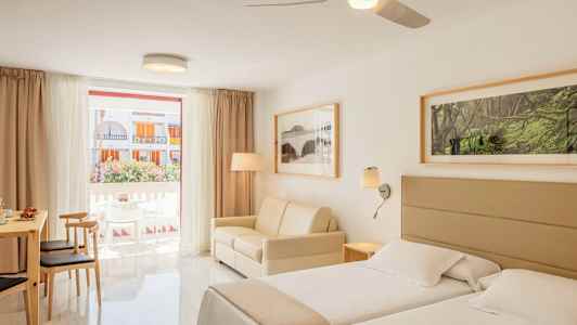 Et lyst hotelværelse med to senge, en sofa og spisebord med stole, samt et stort vindue med udsigt til altanen og bygninger udenfor. (AI-genereret tekst)