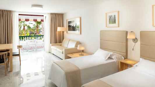 Hotelværelse med to enkeltsenge, en sofa, et bord med stole og et vindue mod en altan. (AI-genereret tekst)