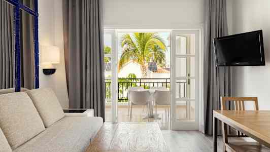 Et opholdsområde med stor sofa, træbord og en TV, der åbner ud mod en balkon med udsigt over tropiske træer og bygninger. (AI-genereret tekst)
