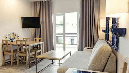 En stue med en grå sofa, et træbord med stole, et vægmonteret TV og glasdøre med gardiner. (AI-genereret tekst)