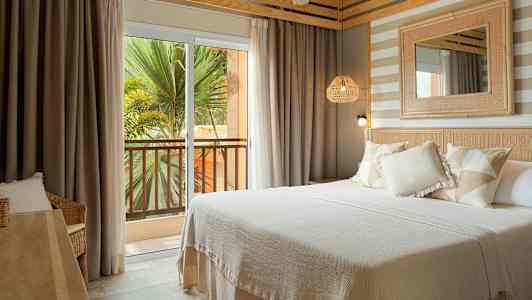 Et hotelværelse med en dobbeltseng, lyse tekstiler og indretningsdetaljer. Udenfor ses en altan med udsigt over grønne områder. (AI-genereret tekst)