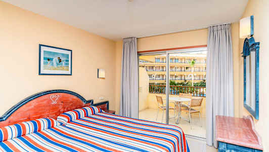 Hotelværelse med to enkeltsenge, stribede blå, røde og hvide sengetæpper, og udsigt til en balkon med møbler. (AI-genereret tekst)