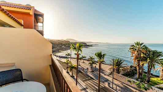 Udsigt fra en balkon mod en palmeomkranset strandpromenade og havet under en klar himmel. (AI-genereret tekst)