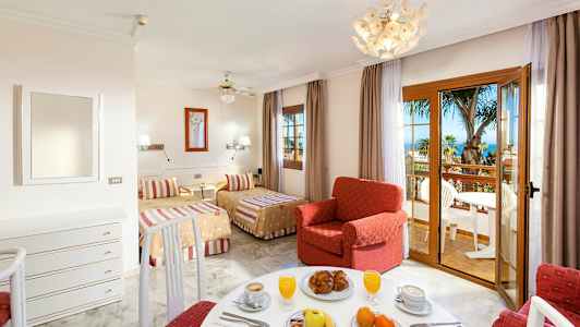 Et hotelværelse med to senge, en rød lænestol og et spiseområde samt en altan med udsigt over palmer og havet. (AI-genereret tekst)