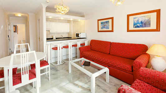 En stue med en sofa og lænestole med rødt tekstilbetræk, et glasbord og et kombineret køkken samt spisestue med hvide møbler. (AI-genereret tekst)