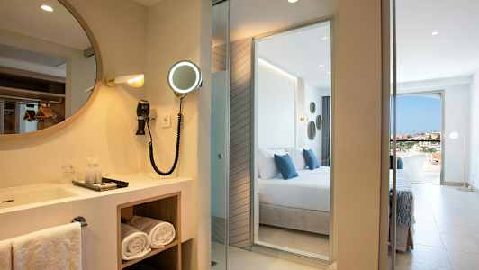 Hotelværelse med et spejl og håndvask ved siden af et soveværelse med udsigt. (AI-genereret tekst)