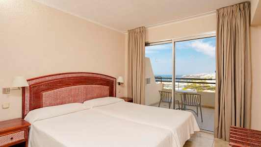 Et hotelværelse med to enkeltsenge, en træhovedgavl og natborde. En glasparti med udsigt over havet og altanmøbler. (AI-genereret tekst)