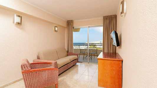 En stue med en sofa, en lænestol, et træbord og et vægmonteret tv. I baggrunden er der en balkon med stole og udsigt over havet. (AI-genereret tekst)