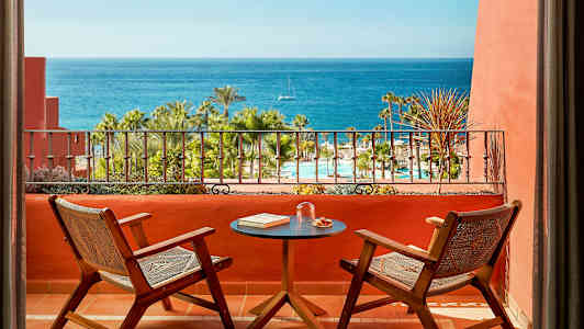 To stole og et bord på en terrasse med udsigt over en pool, palmer og havet. (AI-genereret tekst)