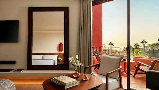 Et elegant hotelværelse med et spejl, åbning mod en møbleret terrasse og havudsigt. (AI-genereret tekst)