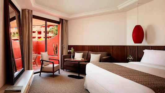 Et hotelværelse med en seng, en sofa, et bord og en lænestol. Værelset har store glasdøre, der fører ud til en balkon. (AI-genereret tekst)