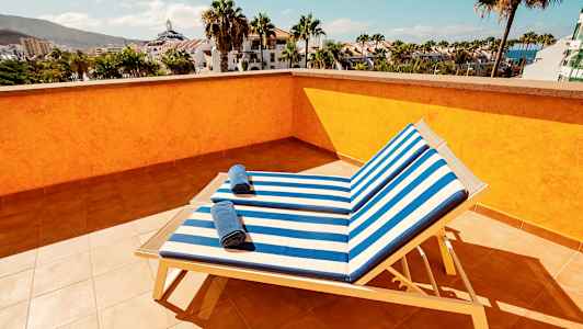Udsigt fra en solterrasse med to solsenge med stribede blå og hvide puder samt håndklæder på, omgivet af palmer og bygninger. (AI-genereret tekst)