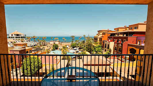 Udsigt fra en altan over et feriested med pools, farverige bygninger og havet i baggrunden. (AI-genereret tekst)