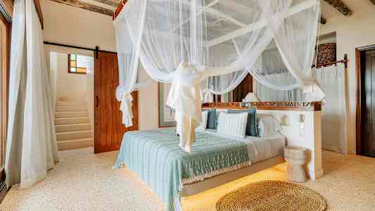 Et soveværelse med en himmelseng med hvidt myggenet, lyse vægge og trægulv. En trappe ses i baggrunden. (AI-genereret tekst)