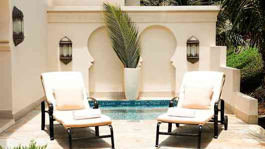 To liggestole med puder og puder står ved en pool foran en lys væg med dekorative udsparinger og grønne planter. (AI-genereret tekst)