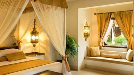 Et elegant soveværelsesinteriør med himmelseng og vinduesplads med guldpuder. (AI-genereret tekst)