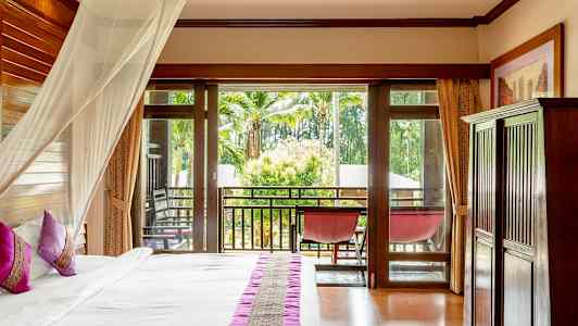 Et hotelværelse med en seng med lyserøde puder og et myggenet, glasdøre fører ud til en balkon med udsigt over palmer og grønt. (AI-genereret tekst)