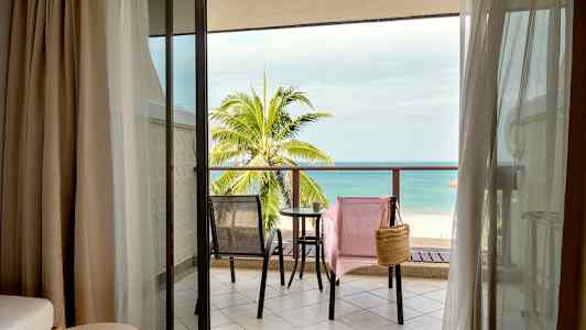 Udsigt fra hotelværelse mod en altan med to stole og et bord, samt en strand og havet i baggrunden. (AI-genereret tekst)