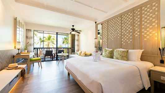 Et hotelværelse med en stor seng dekorret med grønne puder samt et foldet kunstværk forestillende en elefant. Værelset har moderne indretning, et siddeområde og en altan med udsigt til palmer. (AI-genereret tekst)