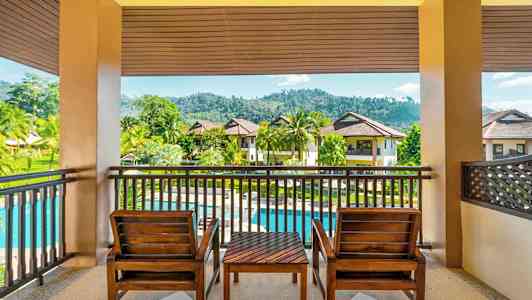 En terrasse med to træstole og et bord med udsigt over en grøn have, en pool og flerbolighuse. (AI-genereret tekst)