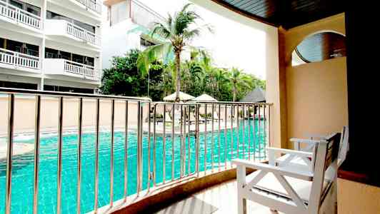 Udsigt fra en altan med hvide stole mod en swimmingpool og omkringliggende bygninger. (AI-genereret tekst)