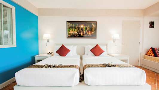 Et hotelværelse med to enkeltsenge, hvidt sengetøj og dekorative røde puder. På væggen hænger et farverigt maleri. (AI-genereret tekst)