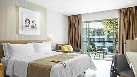 Et hotelværelse med seng, sofa med farverige puder og bord med stol ved et vindue med udsigt til en altan med udsigt over en have. (AI-genereret tekst)