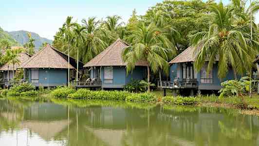Flere blå bungalower med halmtag ligger ved en frodig sø omgivet af palmer. (AI-genereret tekst)