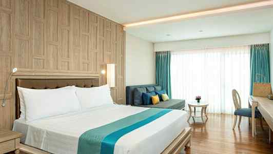 Et hotelværelse med en dobbeltseng, en sofa med puder, et bord og en skrivebordsstol. Rummet har træpaneler på væggene og gulv samt store vinduer med gardiner. (AI-genereret tekst)