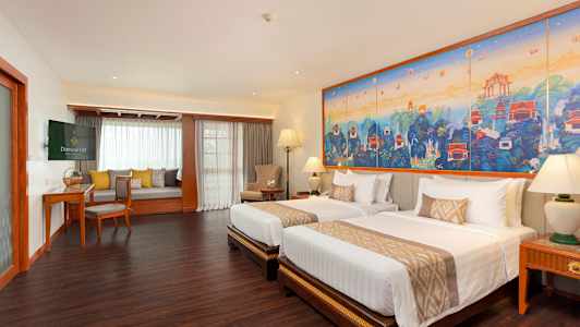 Et hotelværelse med to senge, et stort farverigt kunstværk på væggen og et siddeområde med vindue. (AI-genereret tekst)