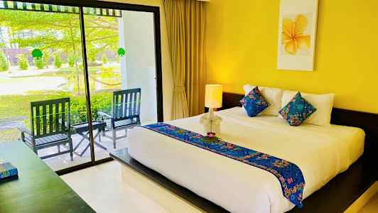 Et hotelværelse med en stor seng dekoreret med farverige puder og et lyst sengetæppe. Sengen er placeret foran en væg med et gult maleri ovenover. En glasdør fører til en terrasse med havemiljø. (AI-genereret tekst)