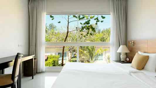 Et hotelværelse med en stor seng, et skrivebord og et vindue, der viser træer og bygninger udenfor. (AI-genereret tekst)