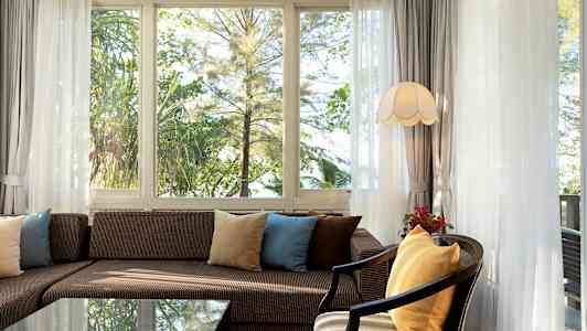 Stue med sofa og puder foran vinduer med udsigt til grønt område. (AI-genereret tekst)