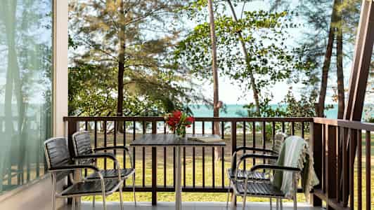 Udsigt fra en veranda med bord og stole, med havet og træer i baggrunden. (AI-genereret tekst)