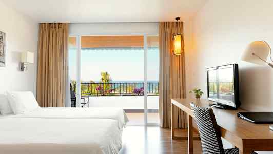 Et hotelværelse med to senge, skrivebord og udsigt til en altan med hav i baggrunden. (AI-genereret tekst)