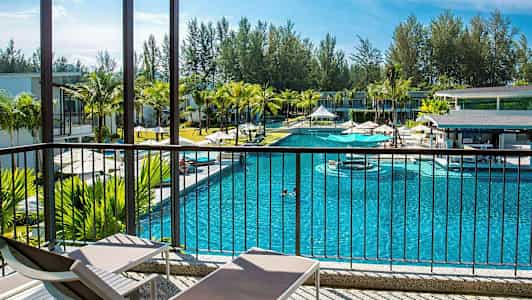 En pool omgivet af bygninger og palmer, set fra en veranda med solsenge. (AI-genereret tekst)