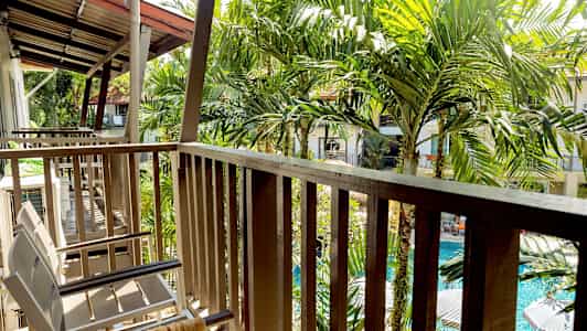 En altan med stole og rækværk, omgivet af grønne palmer og med udsigt over en pool. (AI-genereret tekst)