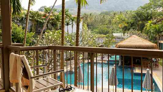 Udsigt fra en altan over en pool omgivet af palmer og grønt, med en stol og et bord i forgrunden. (AI-genereret tekst)