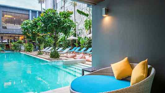 Moderne pool med liggestole og beplantning. En siddeplads med puder i forgrunden. (AI-genereret tekst)